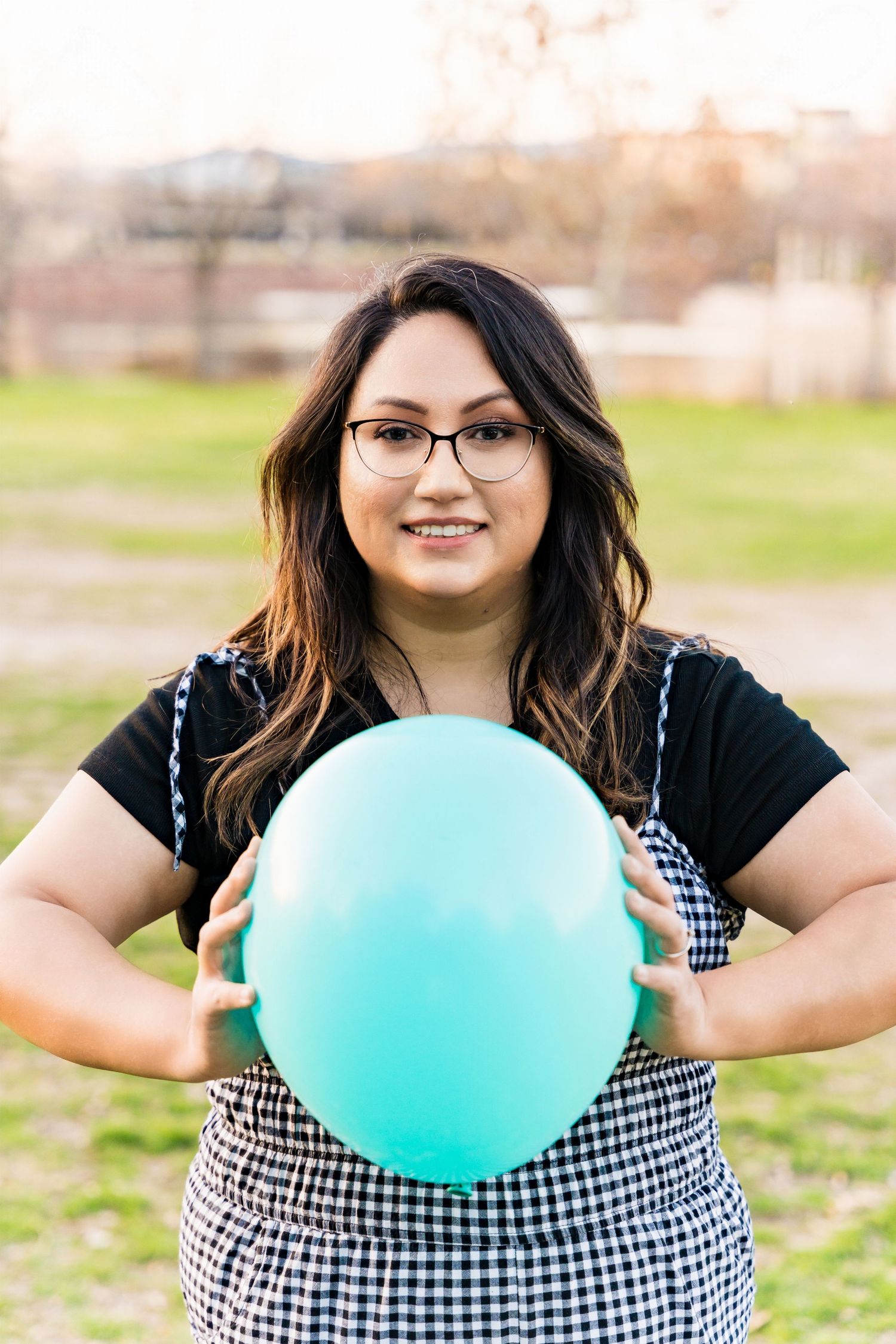 Person holding a large mint green balloon while wearing black and white gingham overalls in a park.