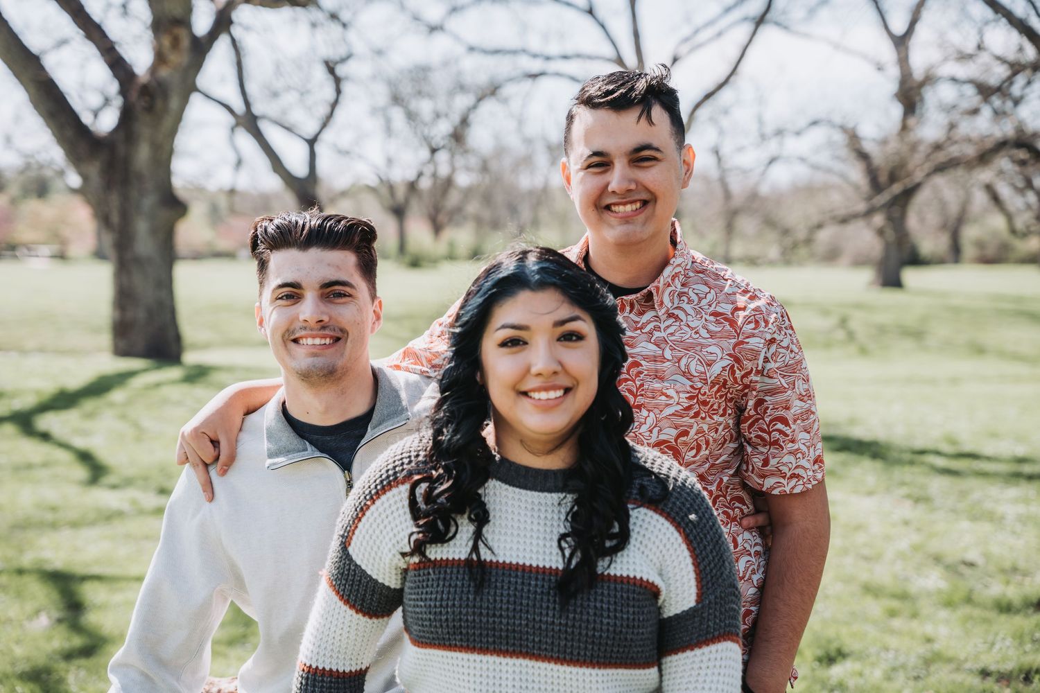 Three friends smile together outdoors with bare trees and green grass in background.