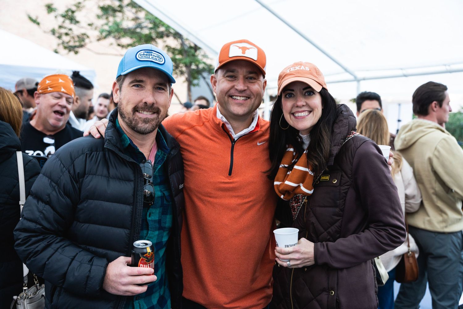 Three friends enjoy drinks together under a tent at an outdoor gathering.