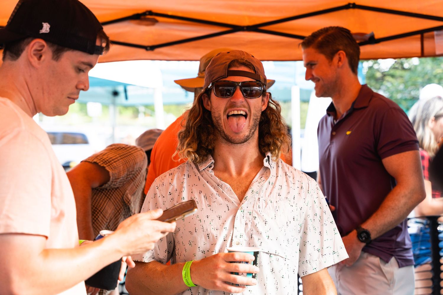 Person in floral shirt and hat enjoying drinks with friends under orange tent.
