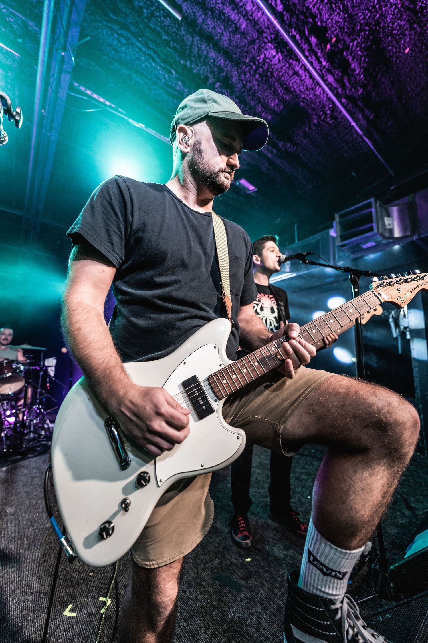 Guitarist in baseball cap plays white electric guitar under turquoise and purple stage lights.