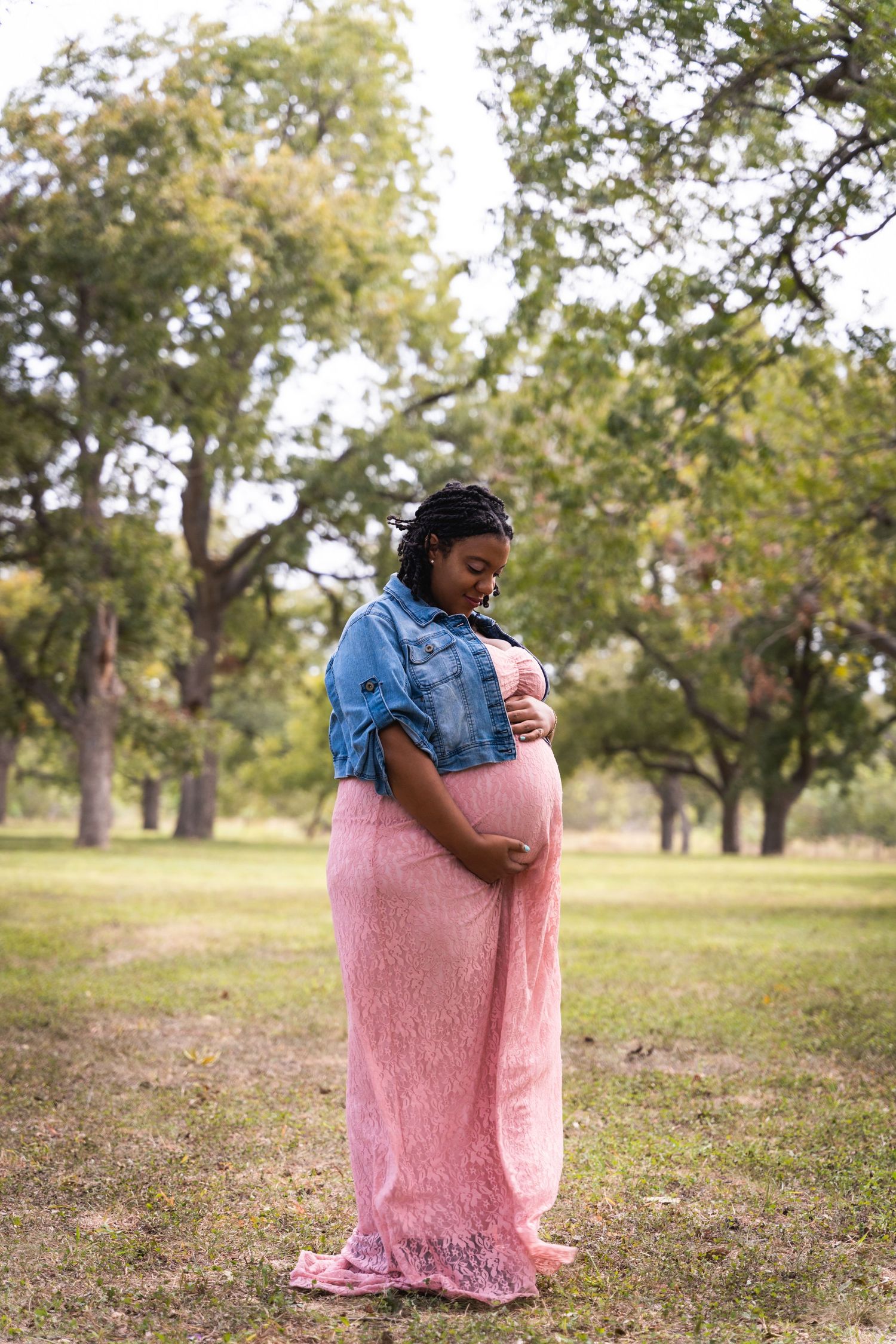 Expectant mother in pink dress and denim jacket stands in sunlit park.
