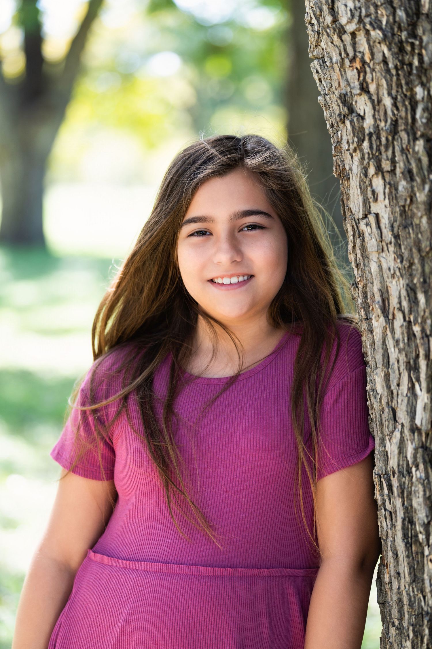 Person in purple dress poses by tree trunk with natural bokeh background and long flowing hair.