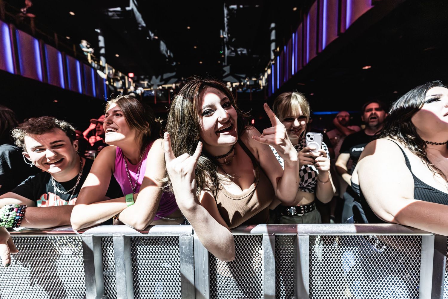 Concert attendees lean on front barrier during live music performance with purple venue lighting.