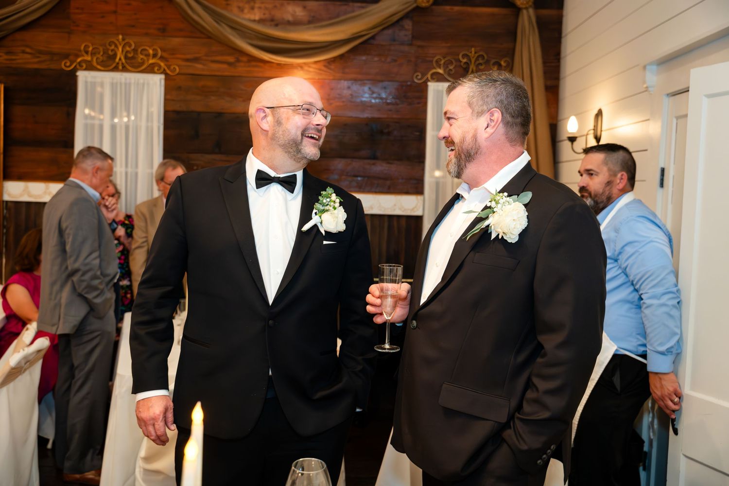 Two people in formal black tuxedos sharing drinks and conversation at an elegant event.