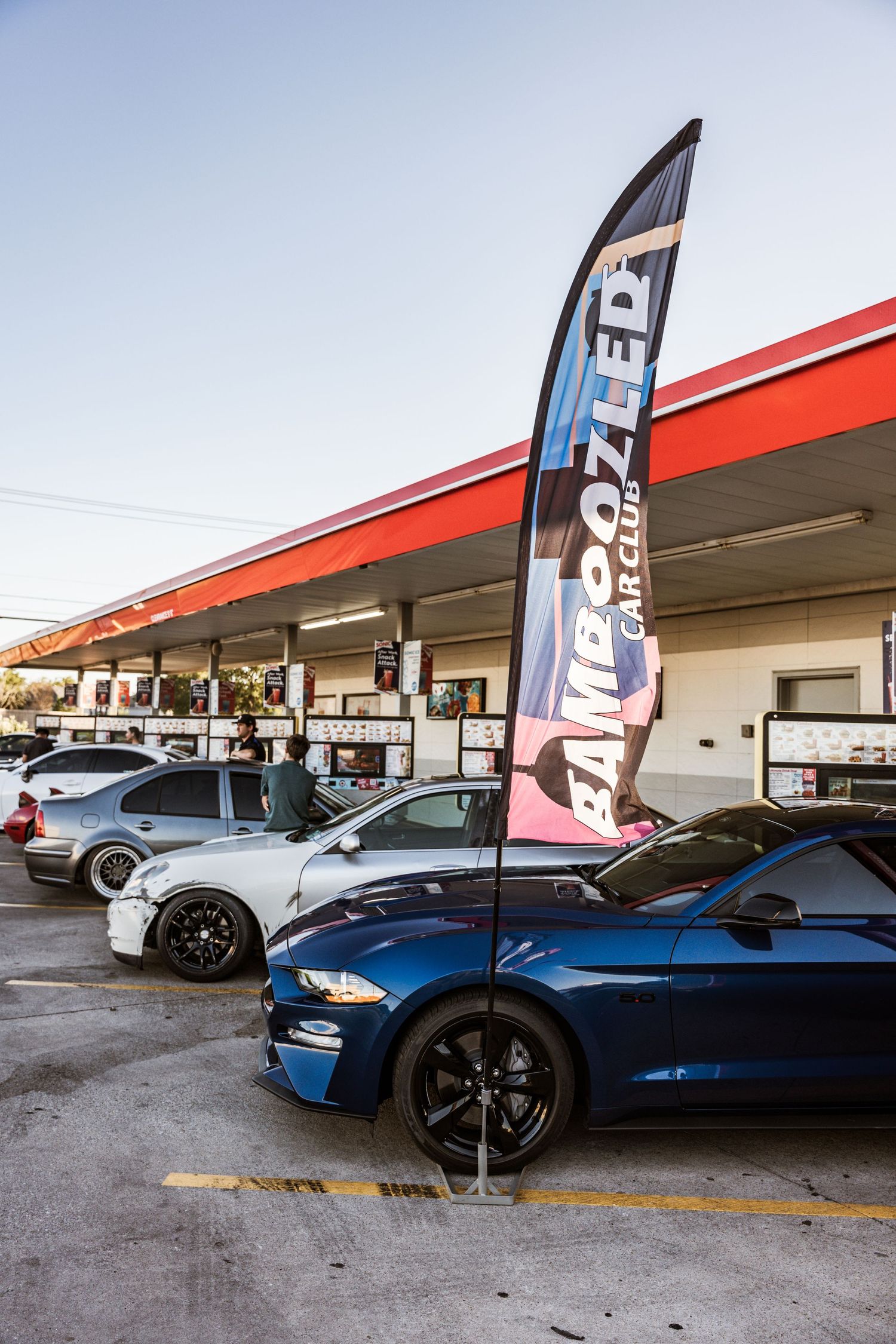 Performance cars lined up at gas station with tall vertical banner flag.
