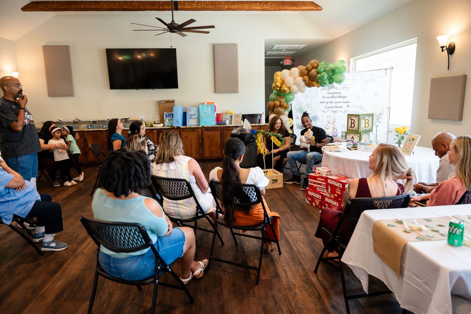 Guests gather in a wood-paneled room decorated for a baby shower with balloons and tables.