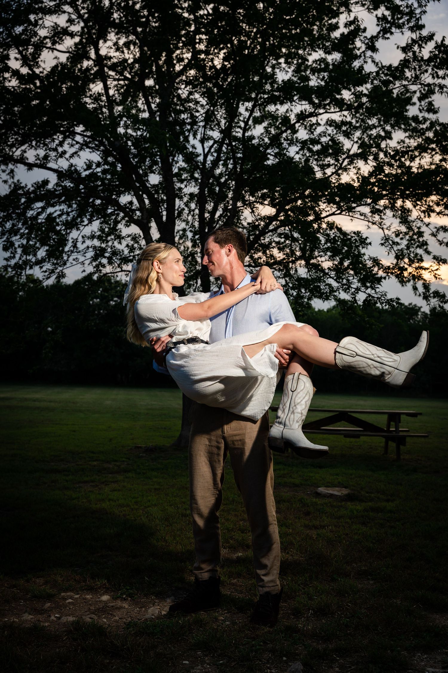 Romantic lift pose captured at sunset with a picnic table and trees in the background.