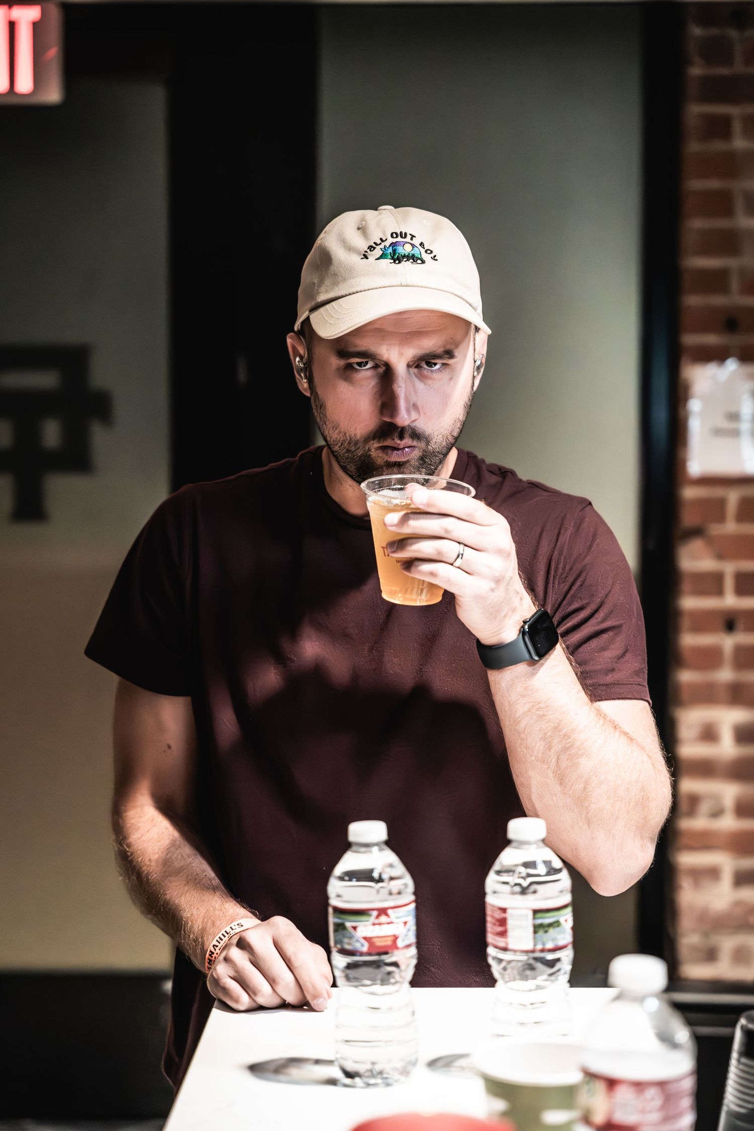 Person wearing a baseball cap and brown t-shirt drinking from a cup with water bottles on the table.