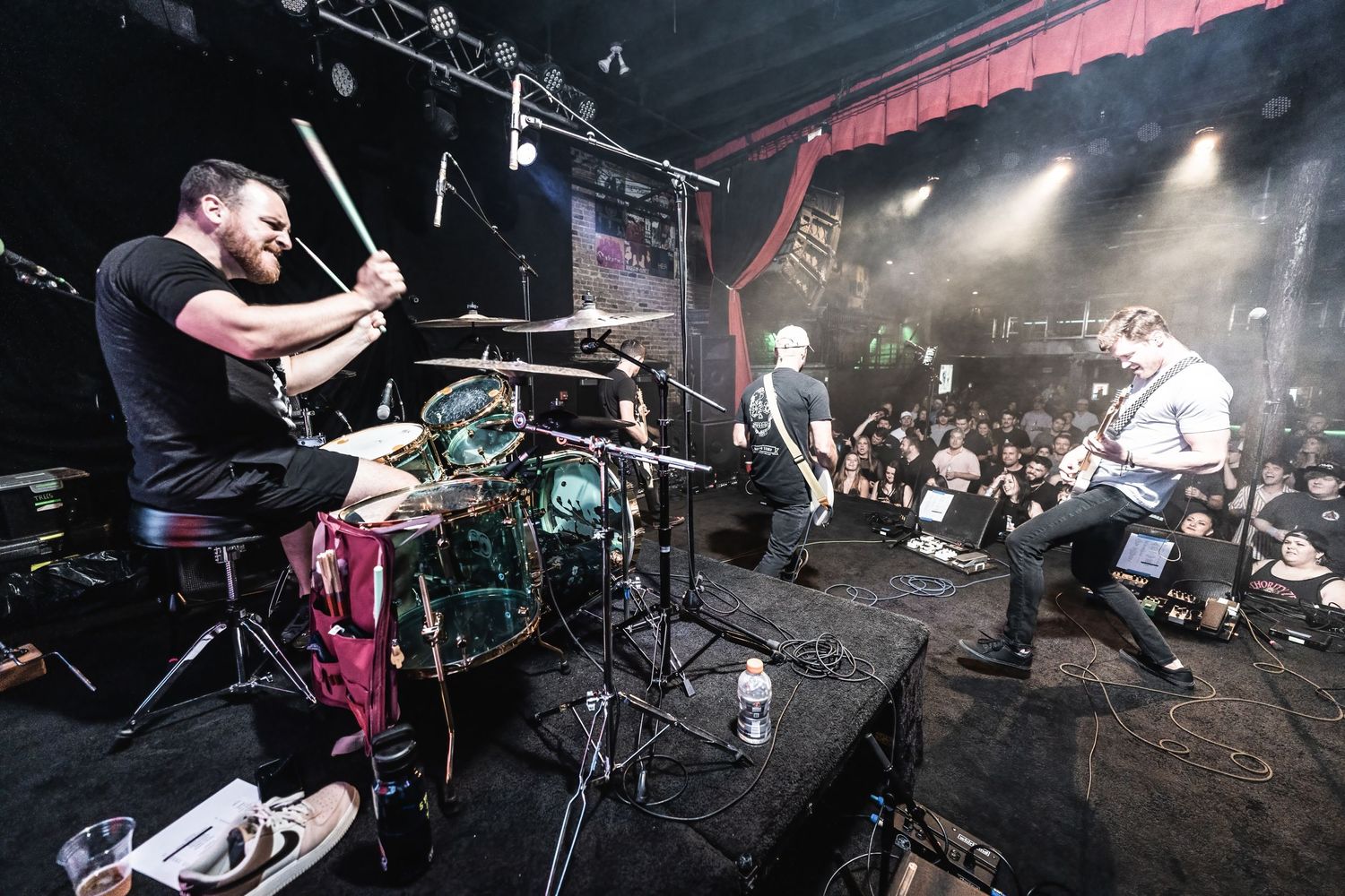 Wide angle shot of full band performing energetically on stage with crowd in foreground.