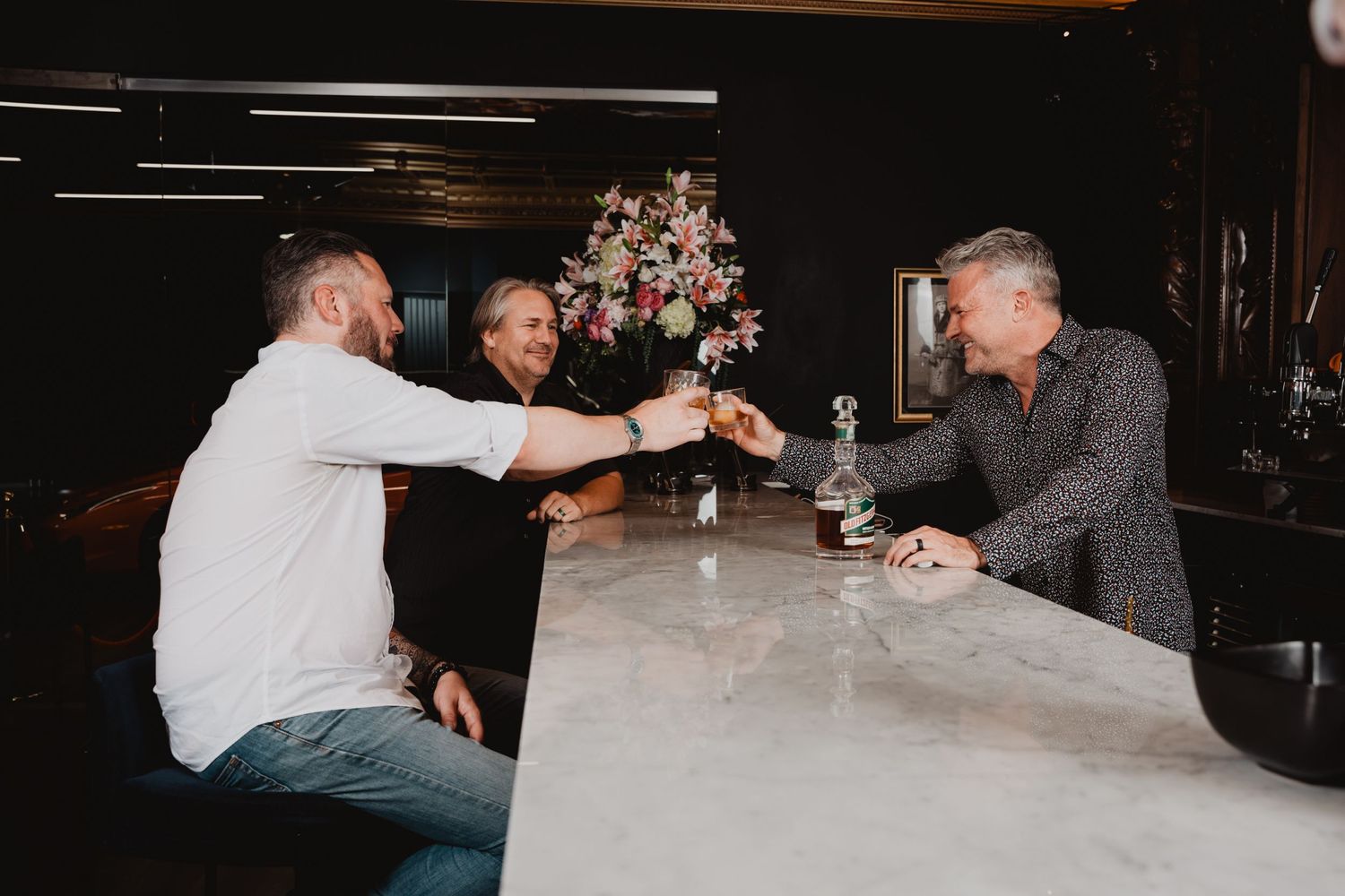 Three people enjoying drinks together at an upscale bar counter with floral arrangements in the background.