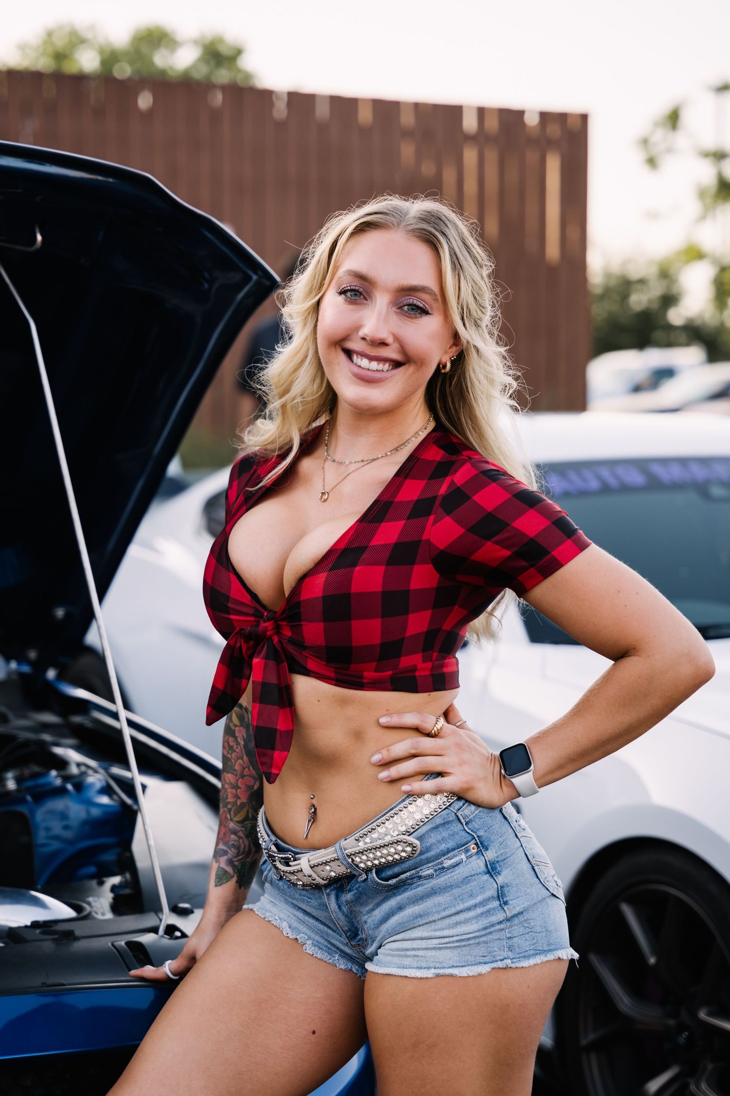 Car enthusiast in plaid shirt and denim shorts poses with modified vehicle at auto show.