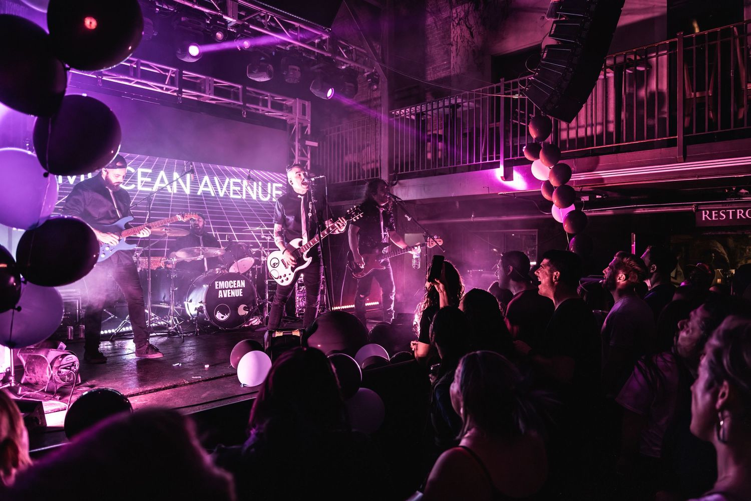 Concert crowd silhouetted against purple stage lighting with Emocean Avenue neon sign and band performing.