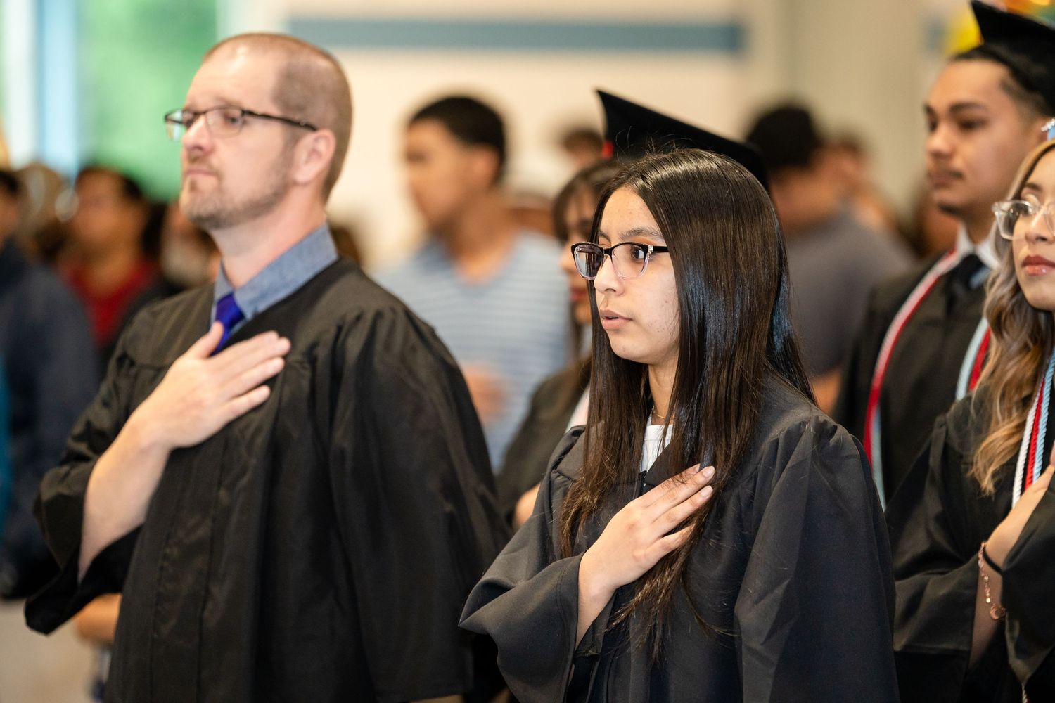 People in academic regalia stand with hands over hearts during a ceremony.