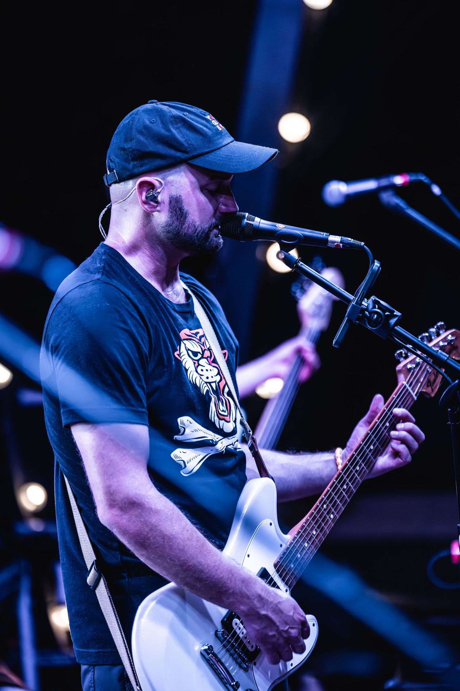 Musician in blue cap playing guitar and singing into microphone under blue stage lighting.