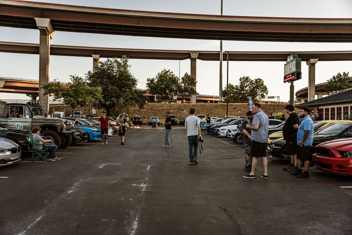 Cars gathered at an outdoor automotive meet under an overpass at dusk.