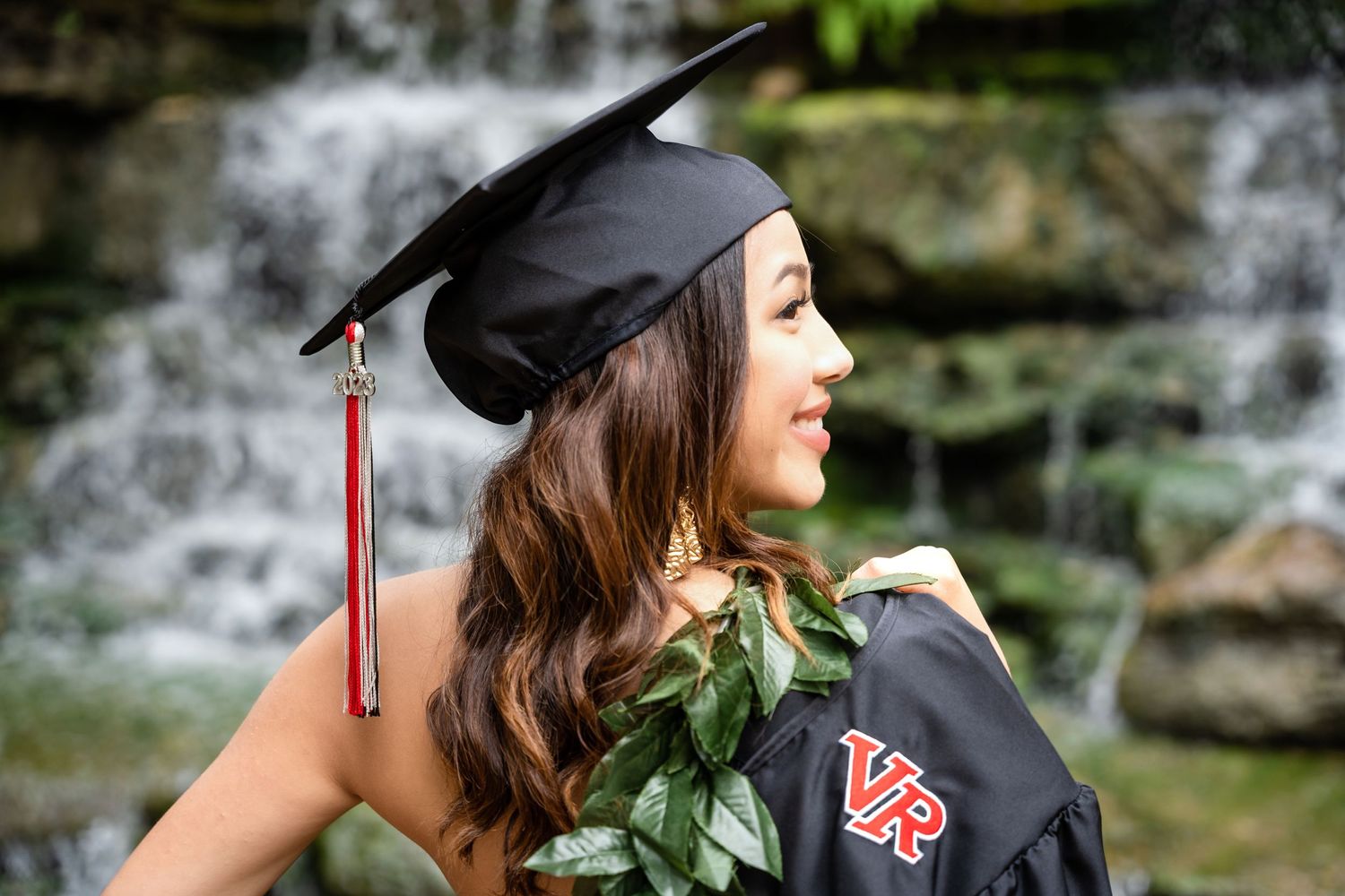 Graduate in black cap and gown with green leaf lei smiling by waterfall.