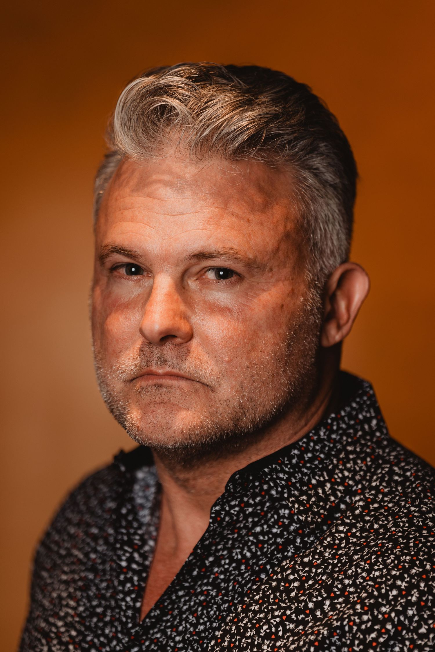 Side-angled portrait of a man with grey hair wearing a patterned shirt against warm orange lighting.