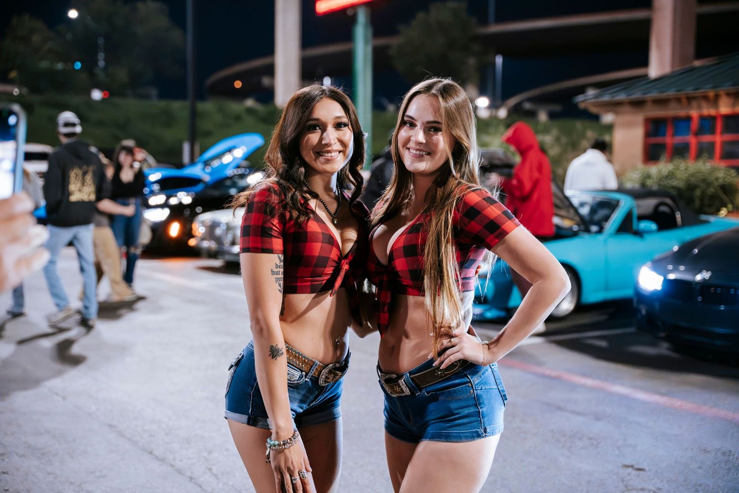 Models in matching red plaid tops at nighttime car show.