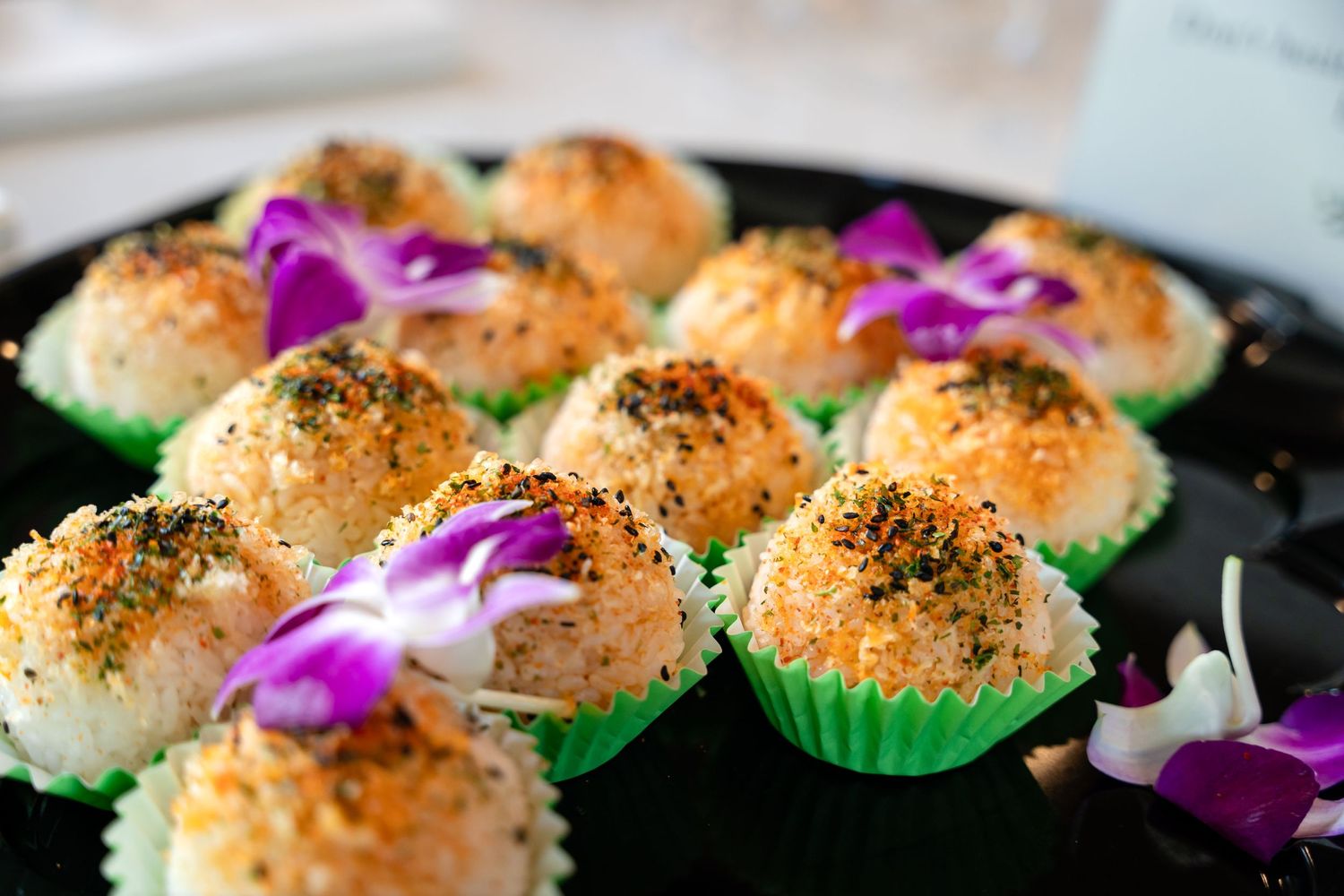 Savory appetizers topped with herbs and garnished with purple flowers served in green paper cups on a black platter.