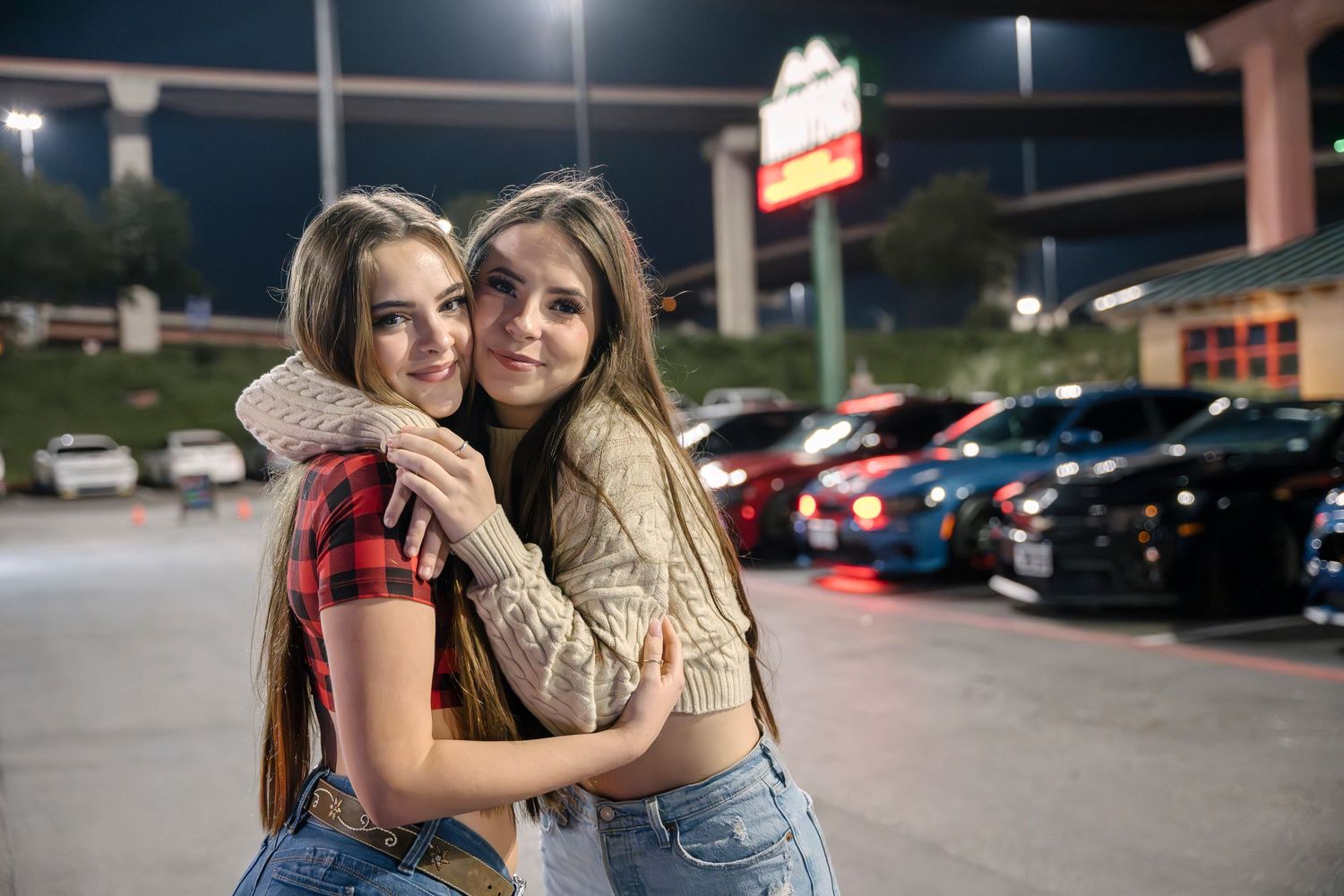 Two friends embracing in casual attire at night in a parking lot with cars in the background.