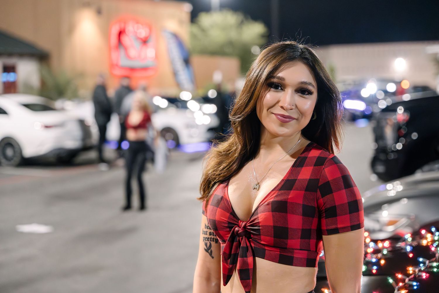 Person in red plaid crop top standing in parking lot at night with cars in background.