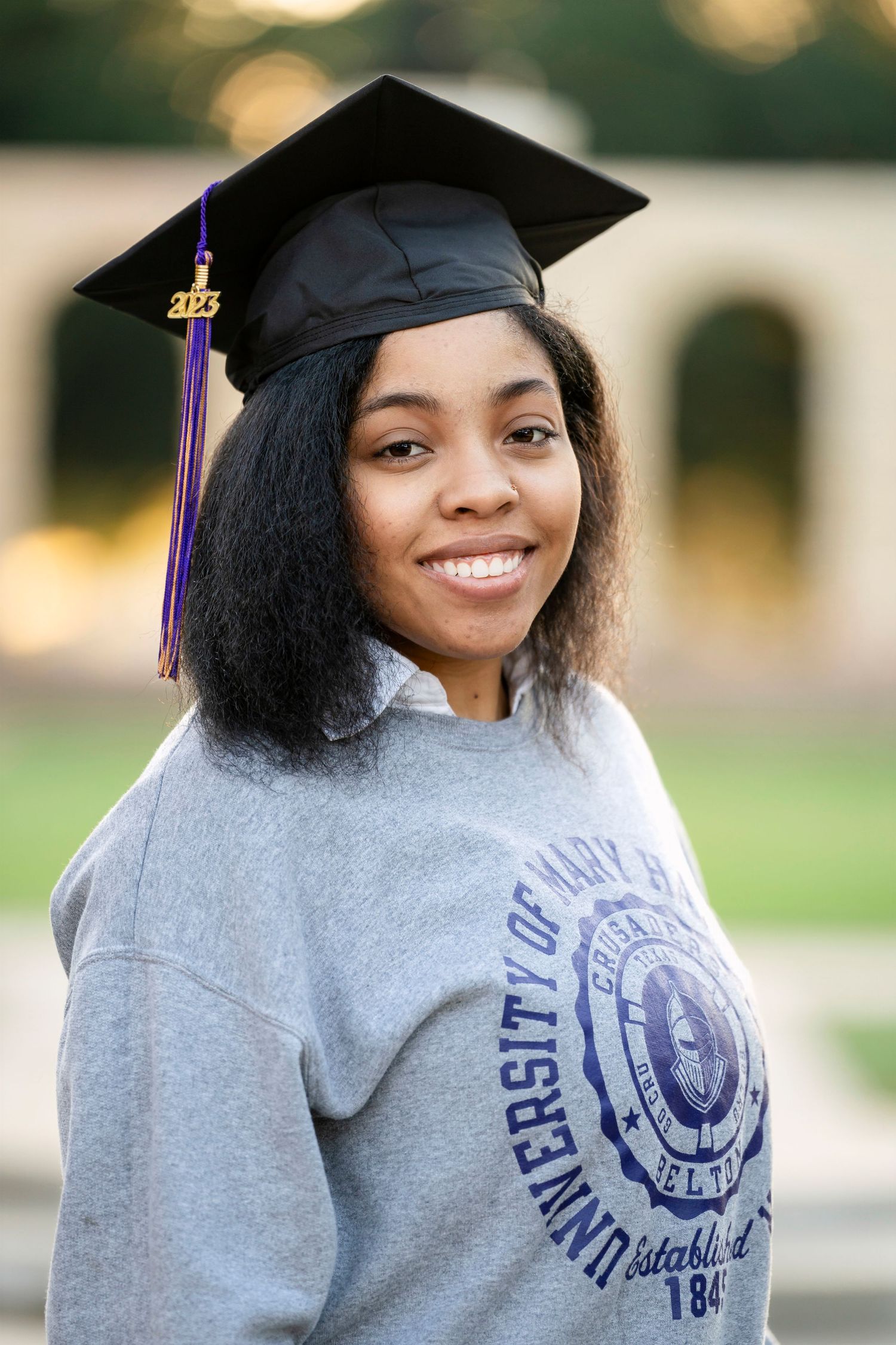A candid graduation portrait featuring a gray university sweatshirt and traditional black mortarboard cap.