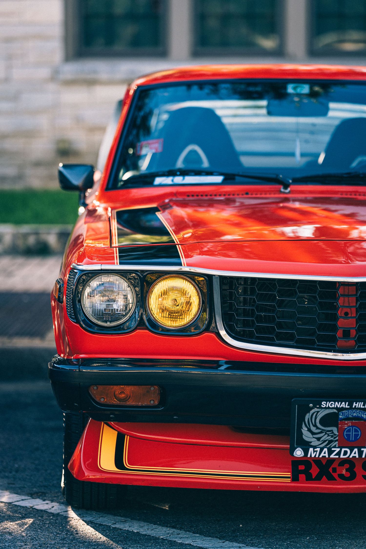 Close up of red classic Mazda RX-3 front end showing yellow fog light and distinctive grille.