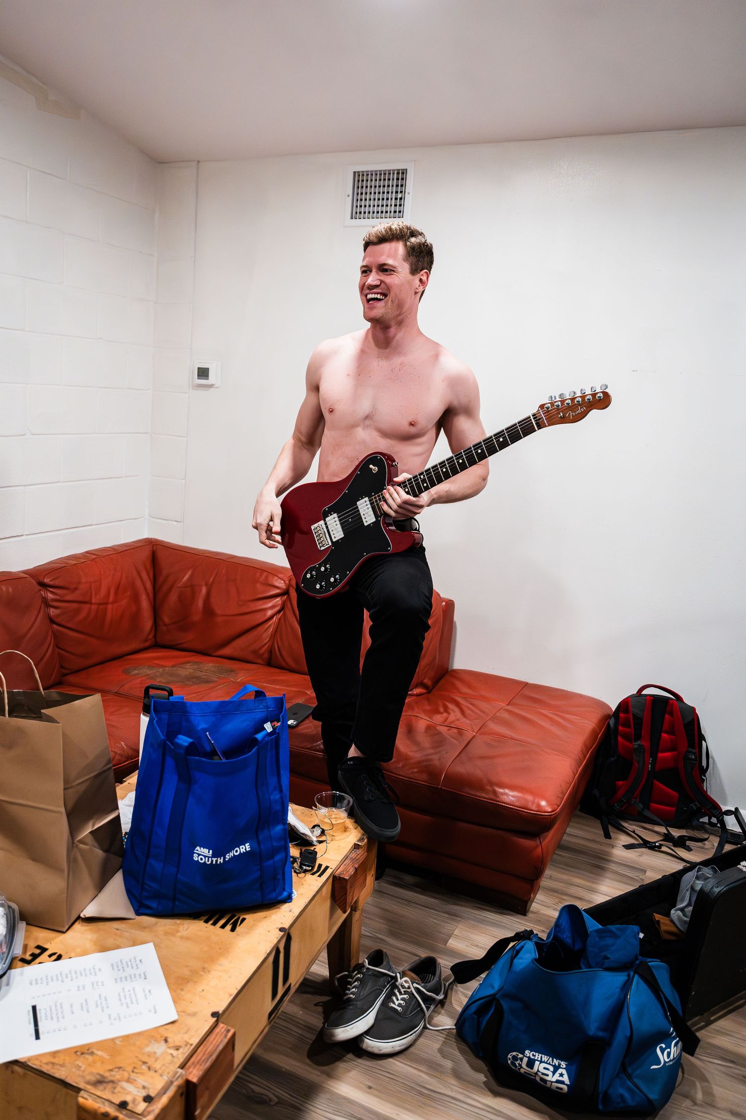 A musician practices with an electric guitar in a backstage dressing room with red furniture and equipment.