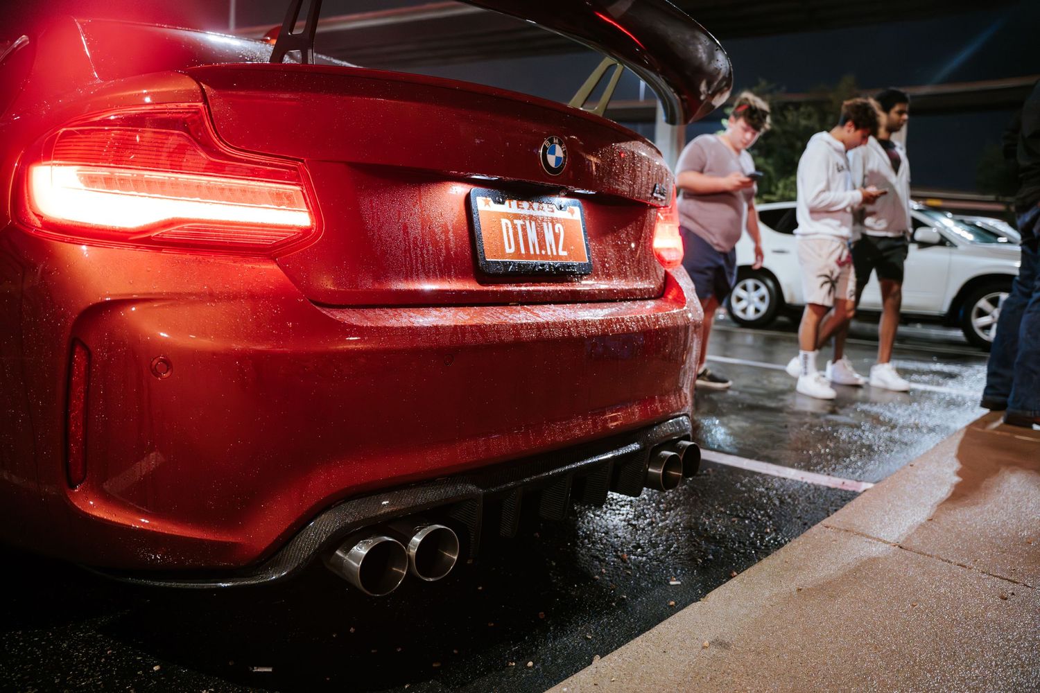 Red BMW M-series rear bumper and taillights with quad exhaust tips in rainy conditions.