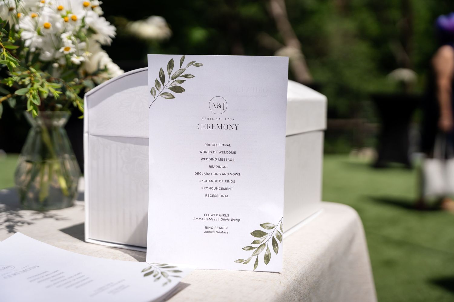 Elegant white wedding program with green botanical designs sits on an outdoor table.