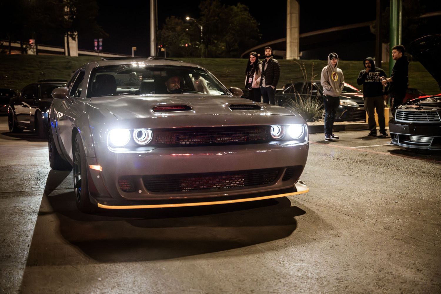 Gray Dodge Challenger with illuminated headlights parked at night car gathering.