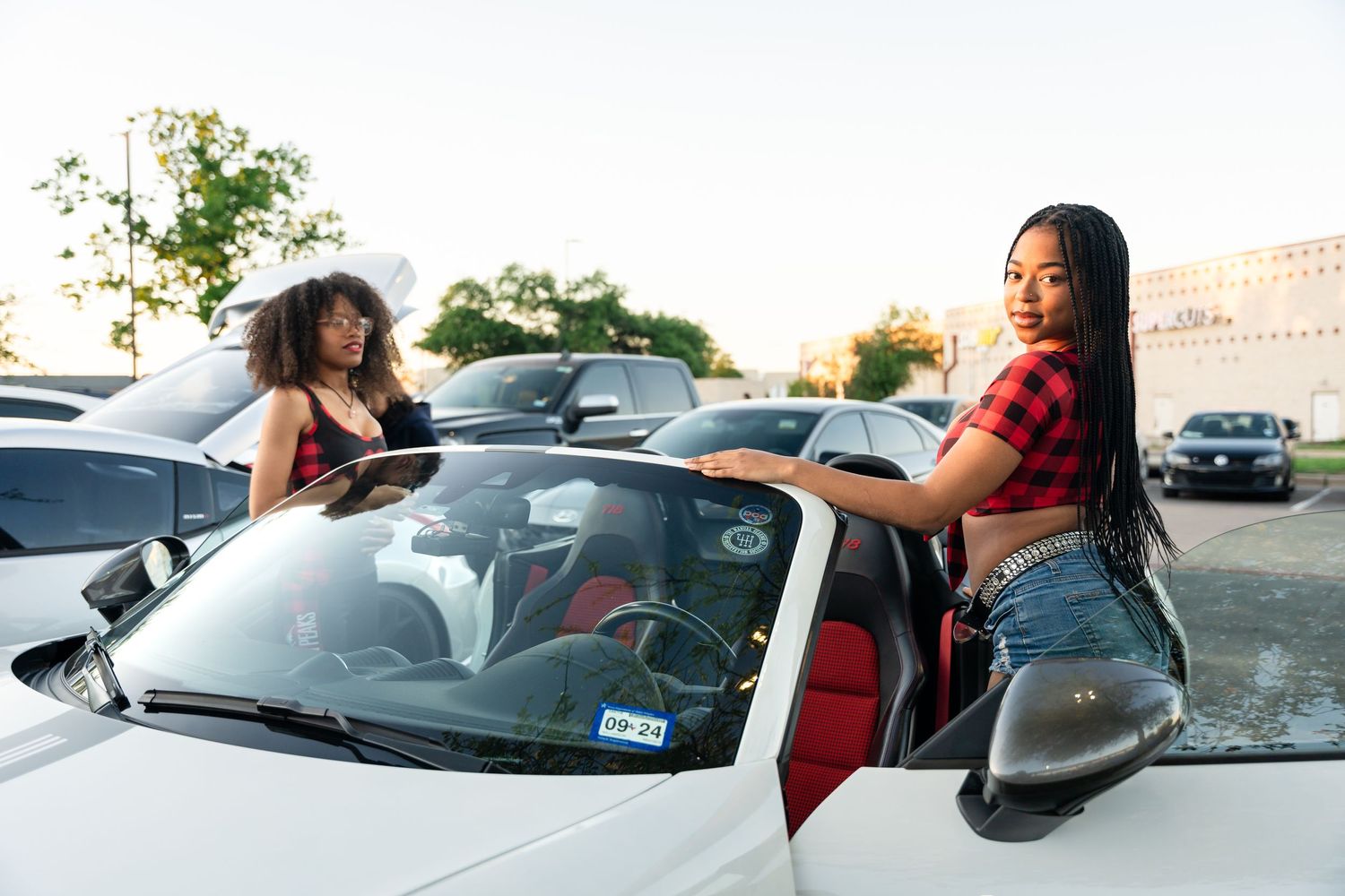 Two people posing with white car in parking lot at sunset.