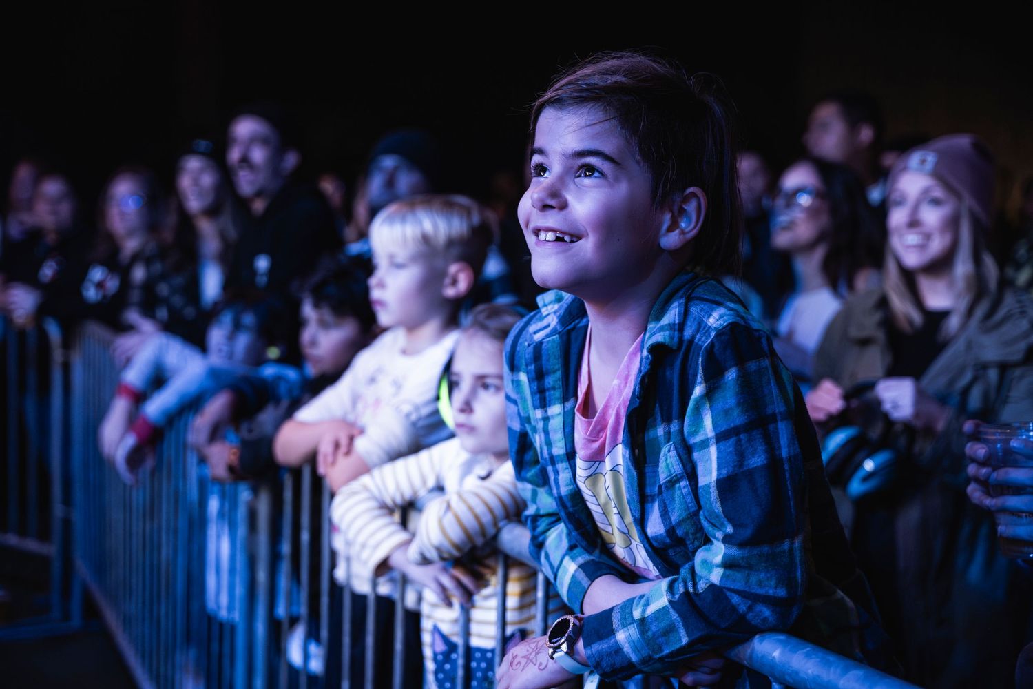 Young concert attendees watch performance from behind security barrier with blue stage lighting.