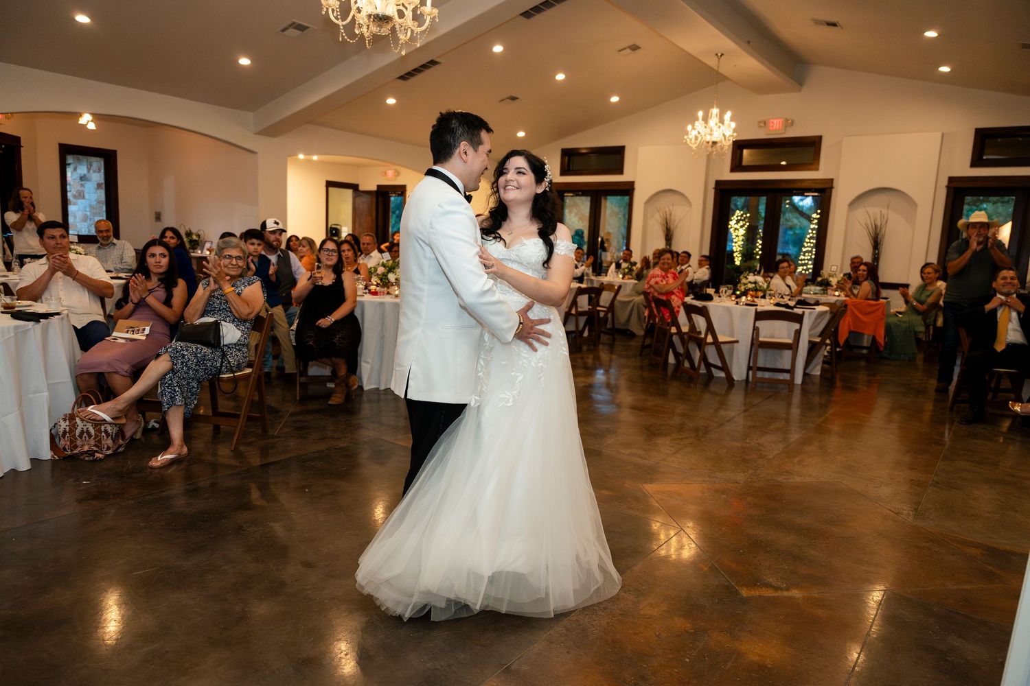 Dancing couple twirls on a polished wooden dance floor in a wedding reception ballroom with chandeliers overhead.