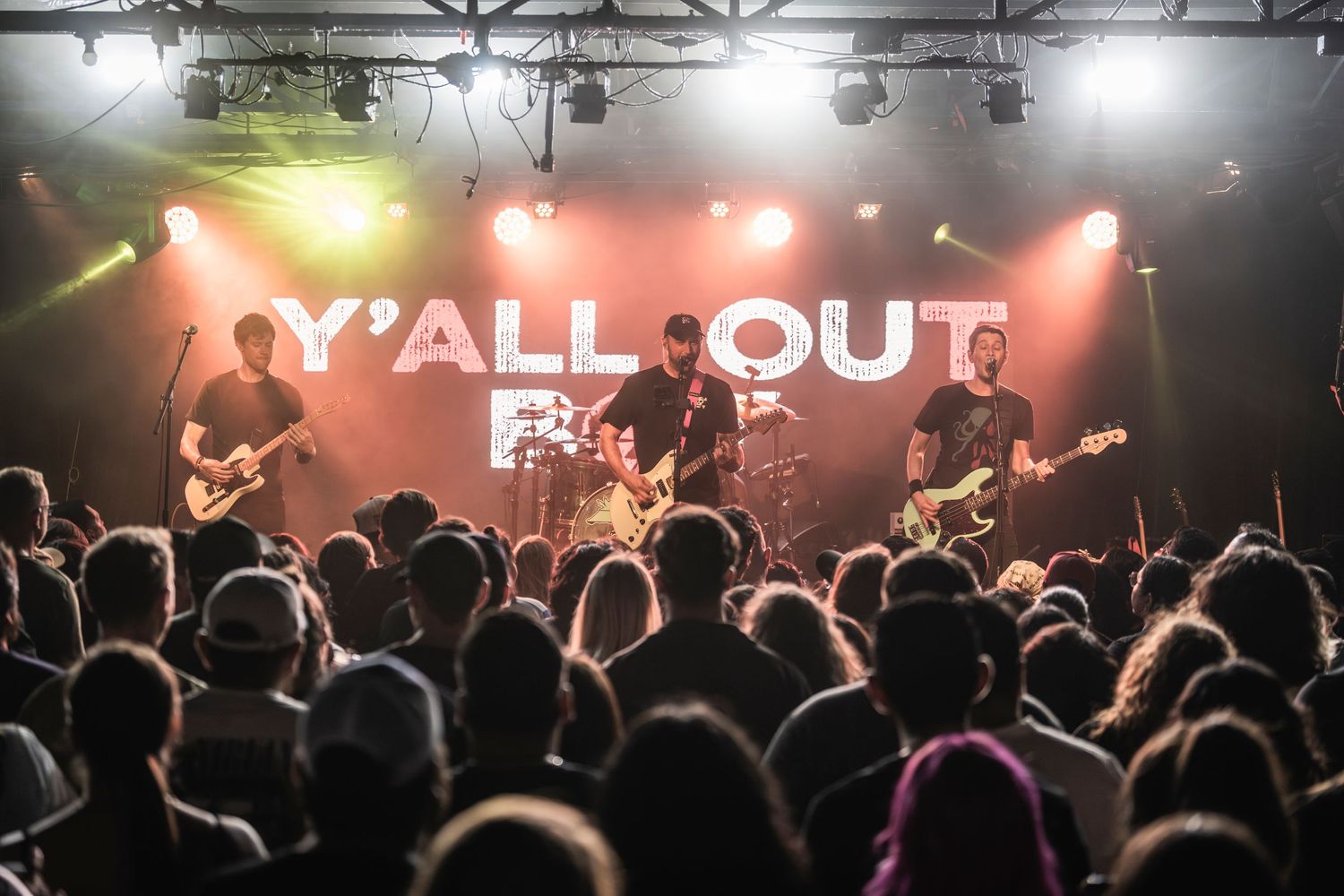 Silhouetted crowd at a rock concert with illuminated stage and neon 'Y'ALL OUT' sign glowing in the background.