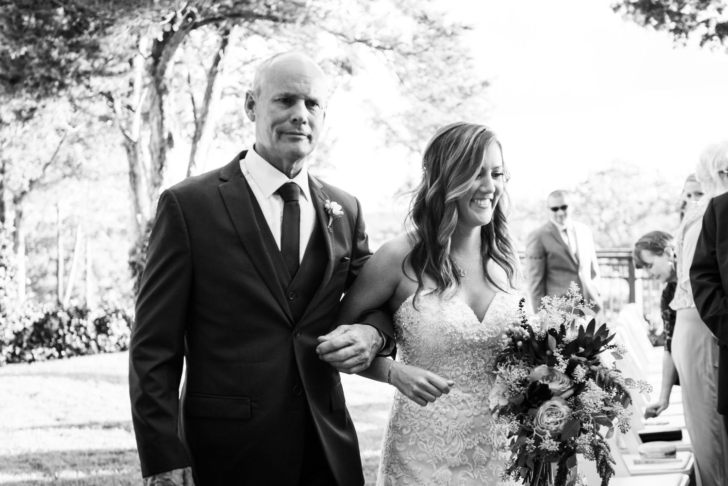 Black and white outdoor wedding processional photo with formal attire and bouquet.