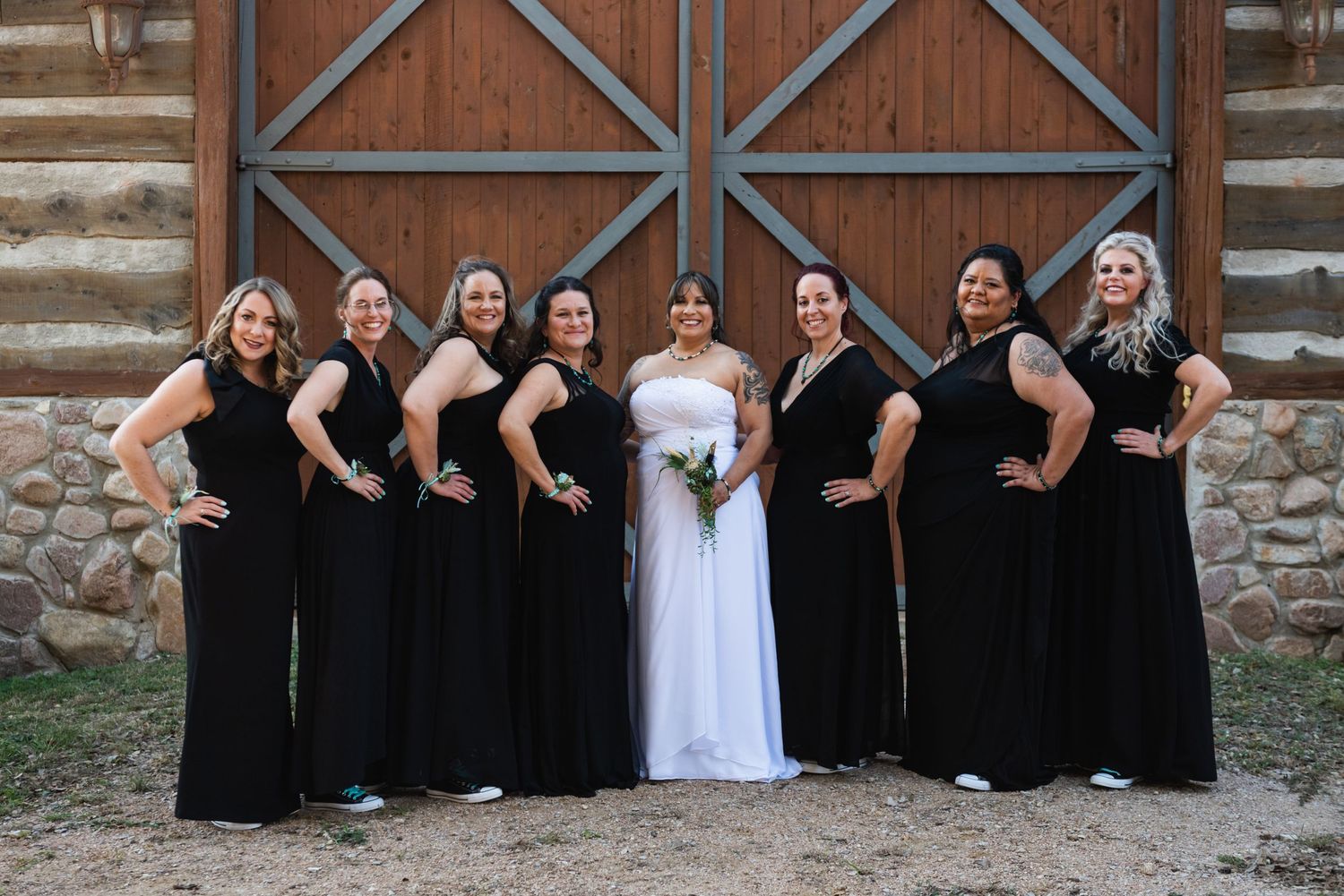 Bridal party in matching black dresses poses with bride against rustic barn wall.
