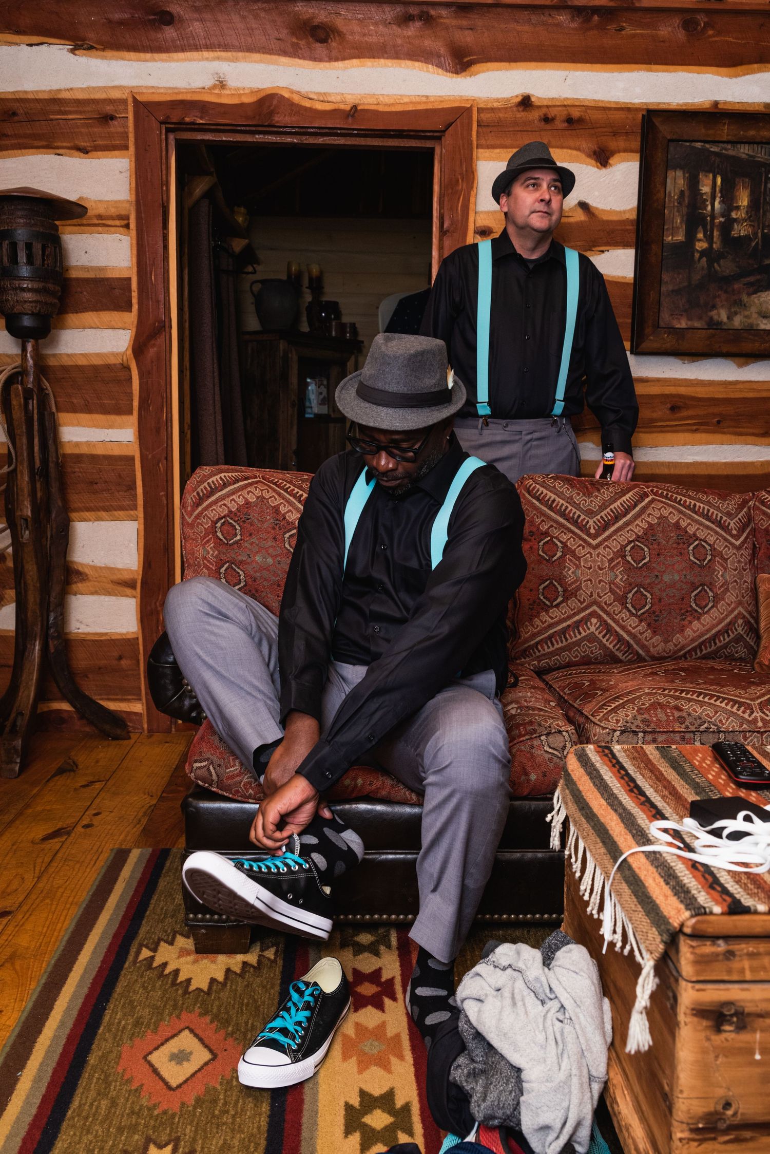Two people in formal wear with suspenders sit in rustic cabin interior.