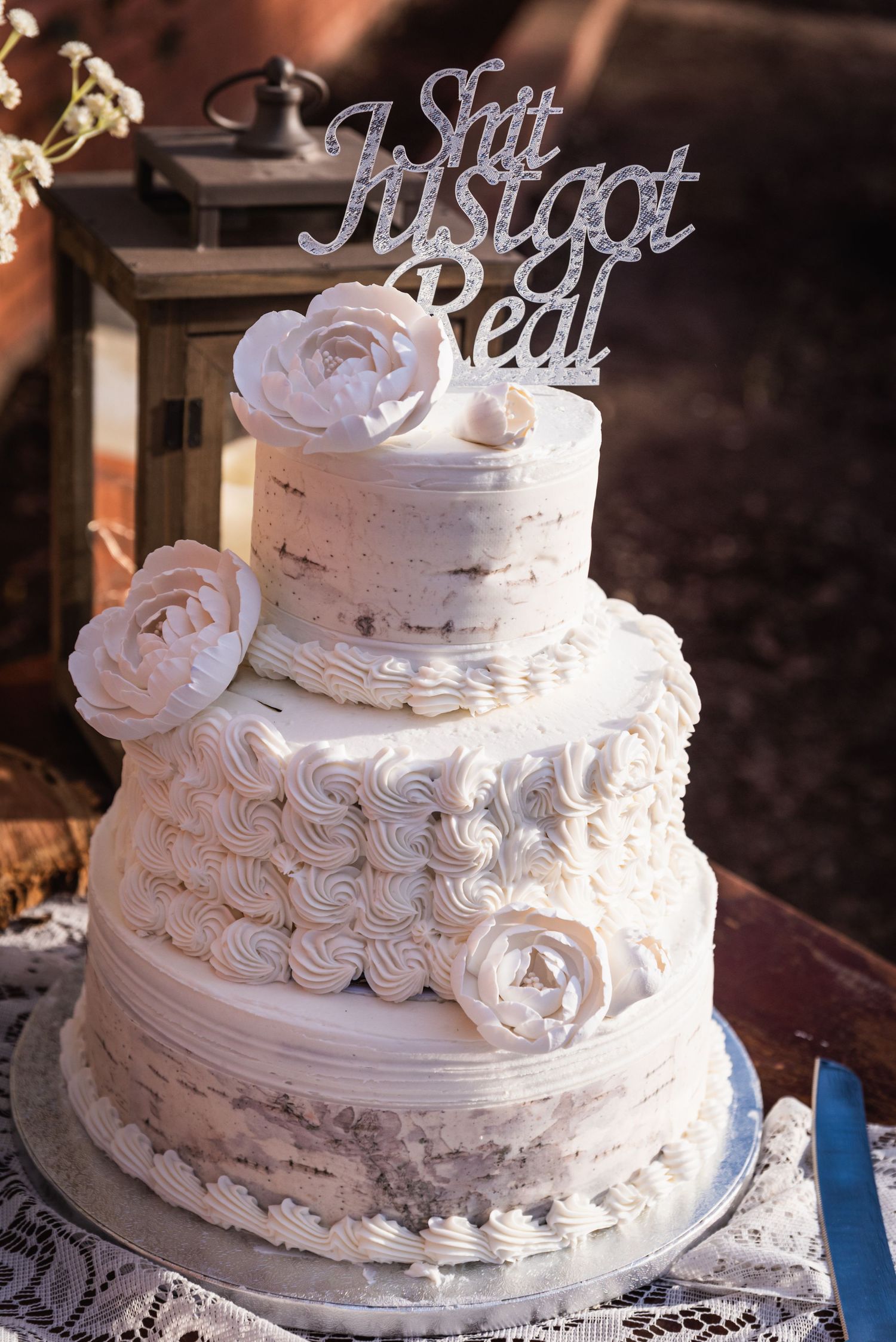 Three-tiered white wedding cake with roses and 'Still Just Got Real' silver topper.
