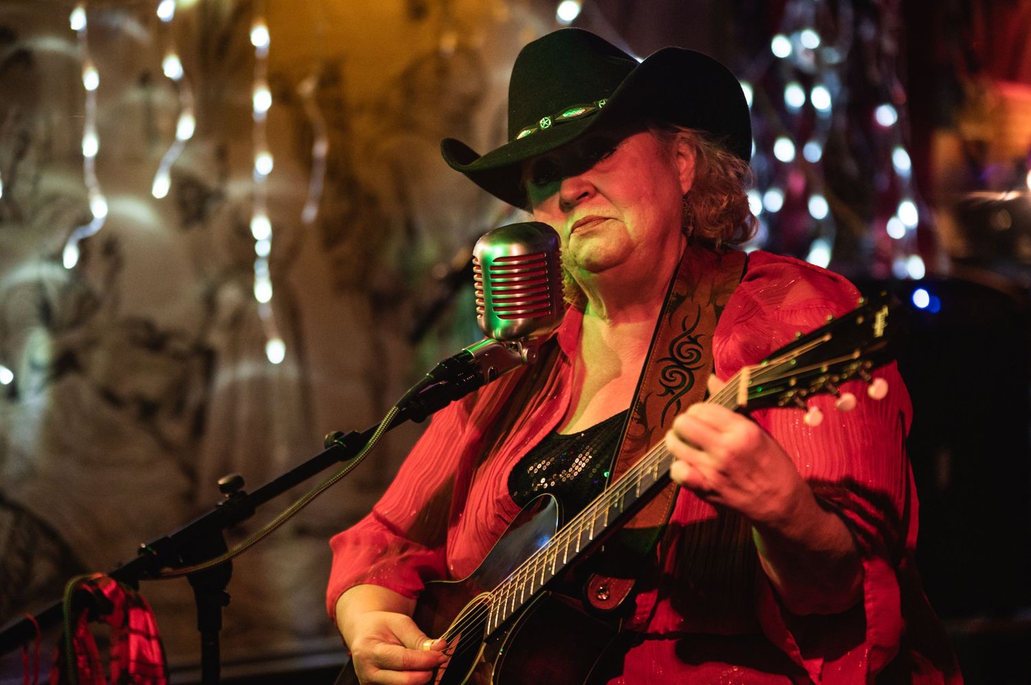 Performer in red jacket and black cowboy hat plays guitar while singing into vintage microphone.