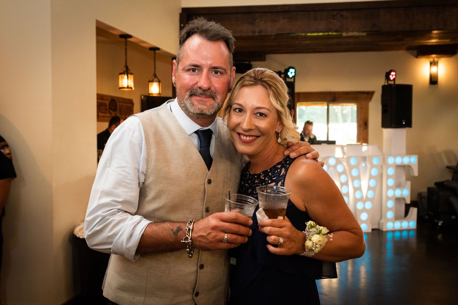 A couple toasts with drinks at a wedding reception venue with exposed wooden beams and ambient lighting.