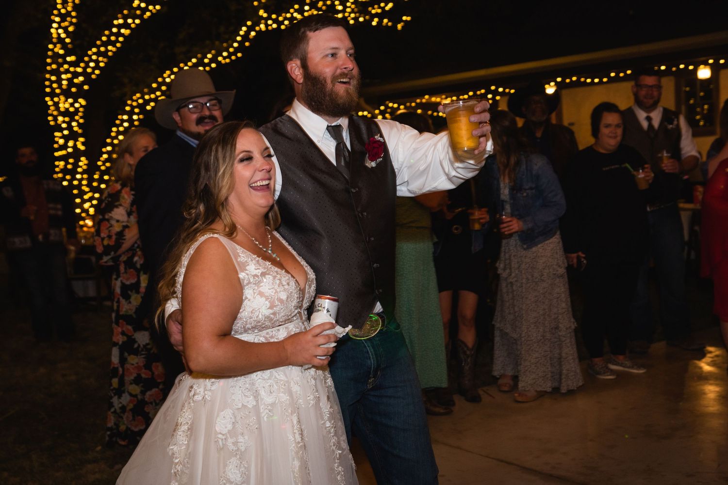 A couple dances joyfully at their wedding reception under twinkling string lights at night.