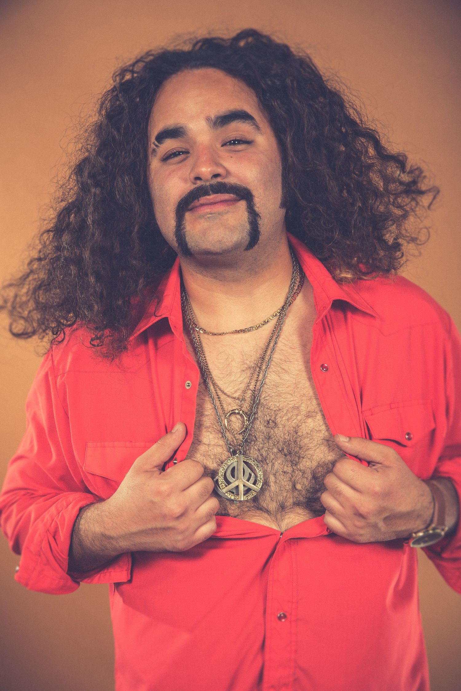 Portrait of someone in red shirt with long curly hair and jewelry against orange background.