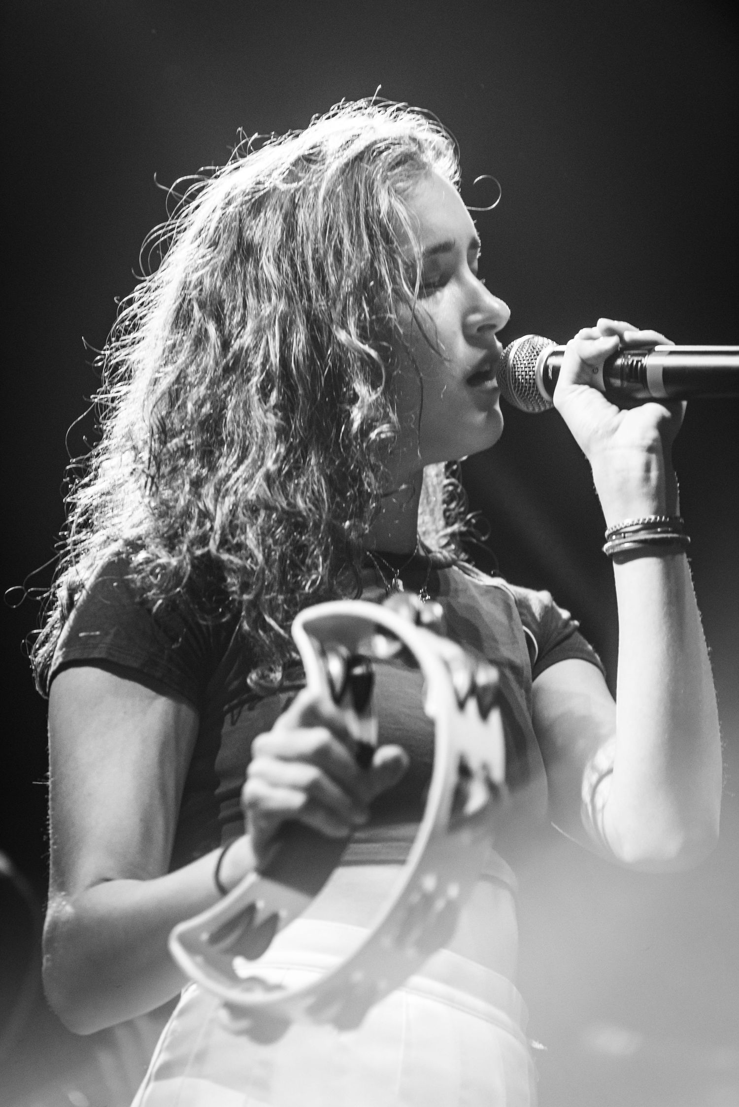Black and white profile shot of performer singing into microphone with curly hair backlit on stage.