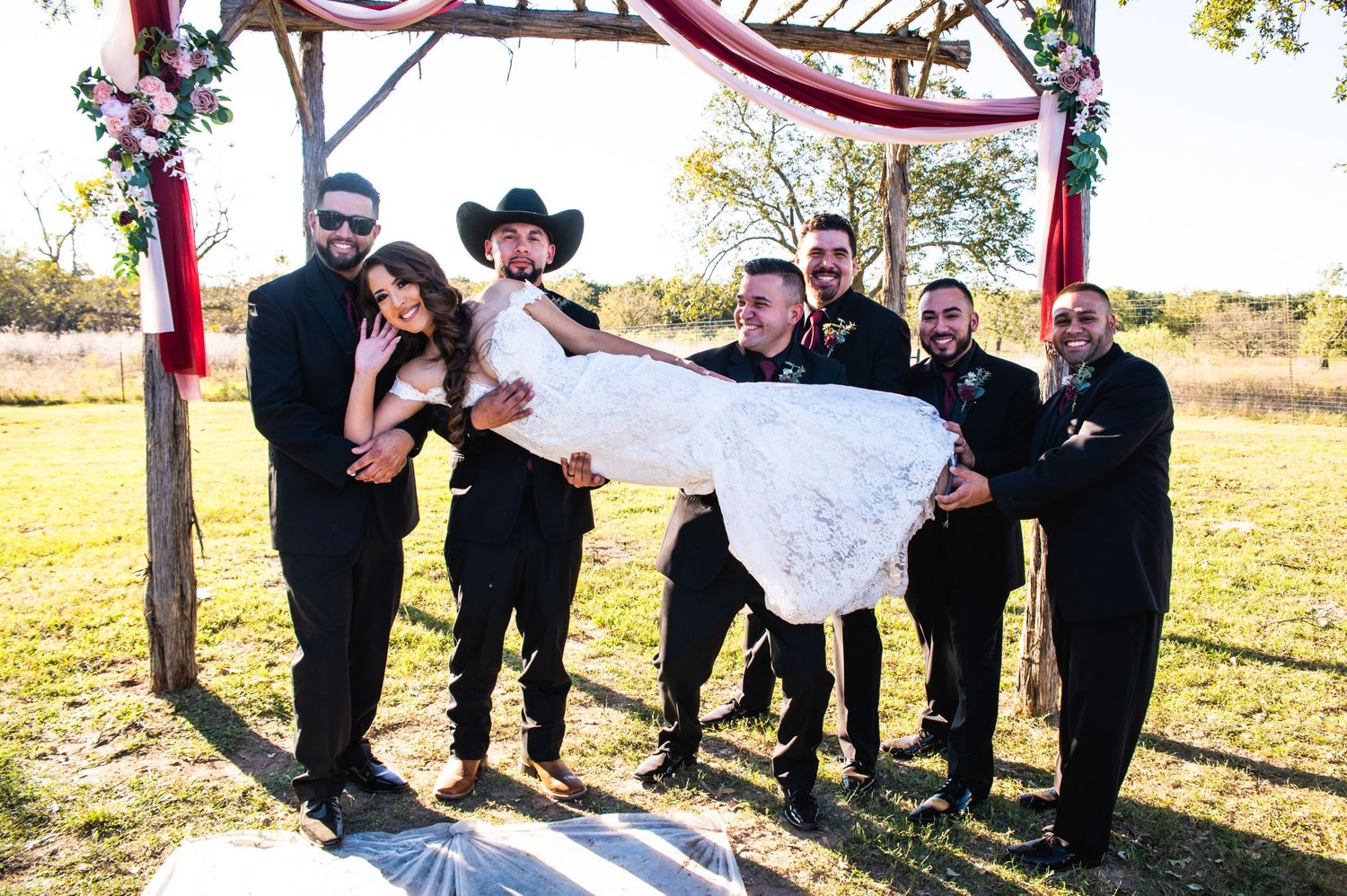 Playful wedding party photo with couple and attendants under rustic wooden arch.