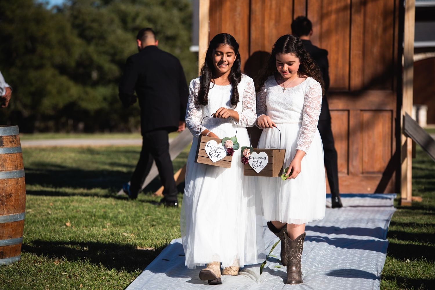 Young flower girls in white dresses walk down an outdoor aisle carrying flower baskets at a rustic barn wedding.