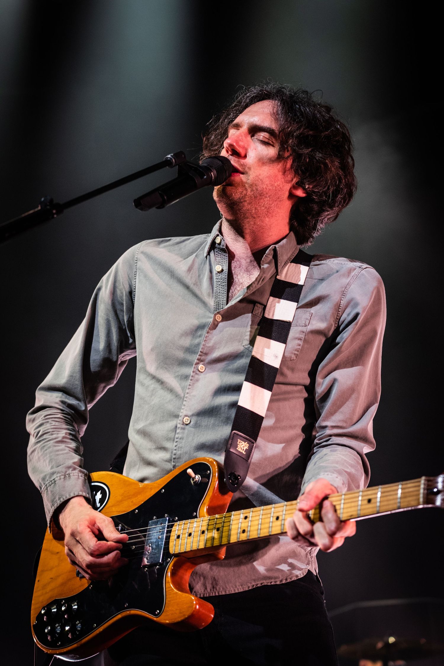 Guitarist plays orange Telecaster guitar while singing on stage with dramatic lighting effects.