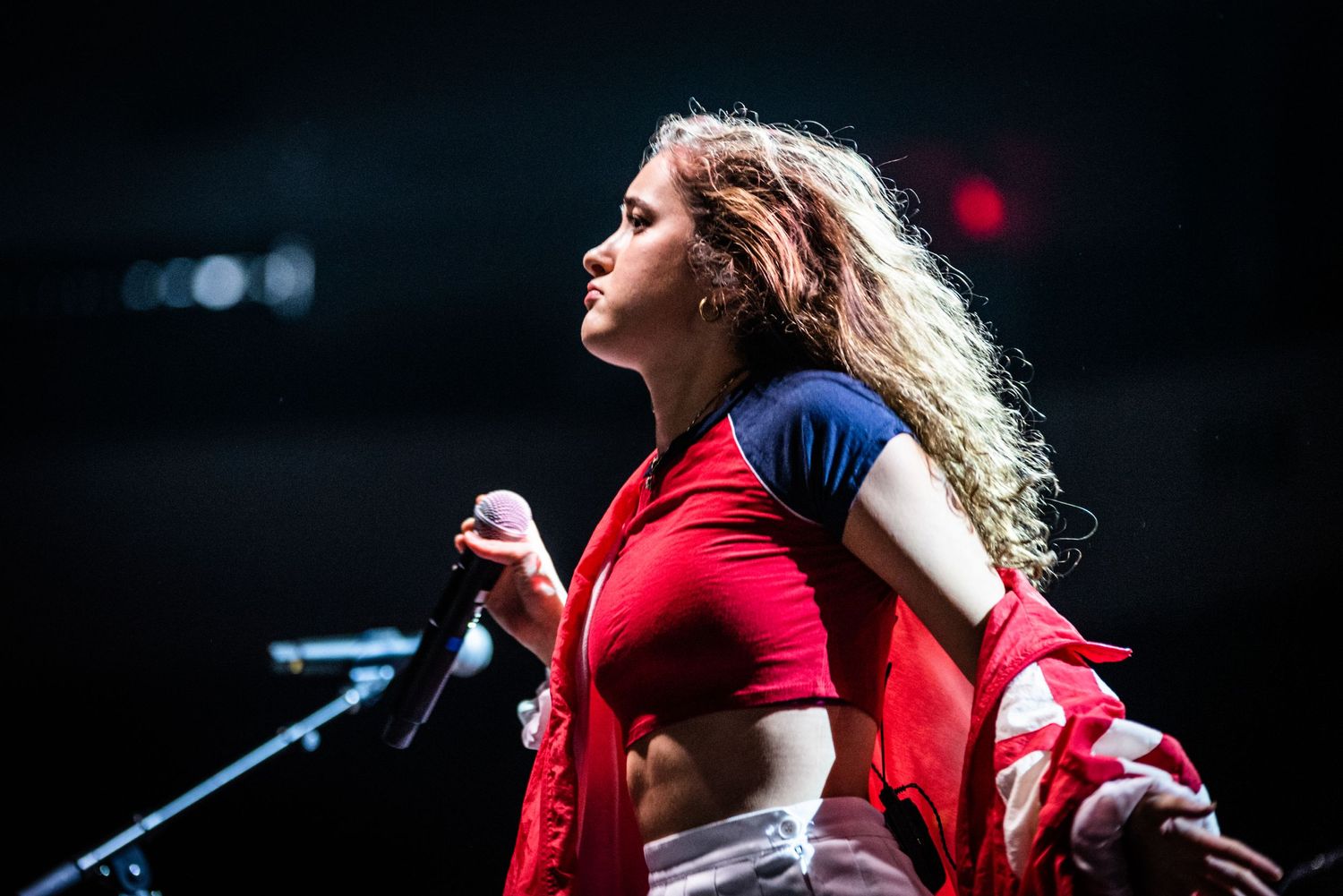 Concert performer in a red and blue outfit dances energetically on stage with dramatic lighting.