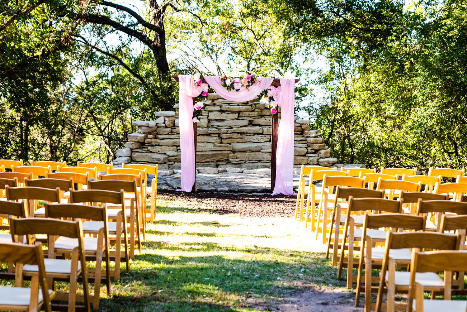 An outdoor wedding ceremony setup with wooden chairs and pink fabric arch against a stone wall.